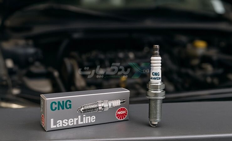 تصویر واقعی شمع مخصوص گاز CNG برند NGK مدل LaserLine در کنار جعبه اصلی روی میز مکانیک با پس‌زمینه موتور خودرو دوگانه‌سوز